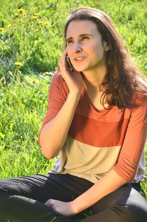 beautiful attractive brunette girl is sitting on a meadow and talking on mobile phoneの写真素材