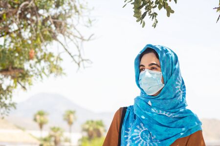 portrait of a young woman with a face mask and a blue hijab surrounded by trees and leaves looking...