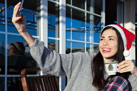 A beautiful young woman smiling holding a cup and taking a selfie with her mobile phone while wearing a Santa Claus hat at Christmasの写真素材