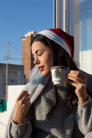 A beautiful woman drinking a Christmas mug while wearing a facial mask, a winter blanket and a santa...
