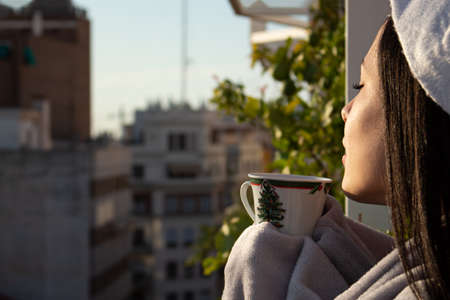A beautiful woman drinking a Christmas mug while wearing a facial mask, a winter blanket and a santa claus hatの写真素材