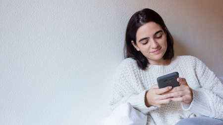 A woman indoor portrait. Young beautiful using her mobile phone. woman in warm knitted clothes at...