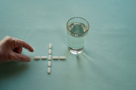 White tablets form a cross and a glass of water on a light, surface. A hand in a surgical glove reaches for a tablet. The concept of selective, discriminating medication therapy.の写真素材