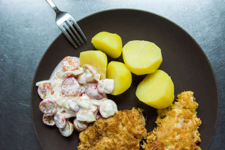 Pork chop, potatoes and tomatoes with sour cream on a brown plate, beside it a fork, an aluminum...