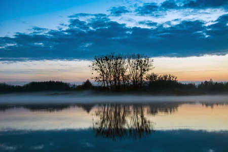 Trees on the edge of a calm lake reflecting in the water, evening fog and clouds in the skyの写真素材