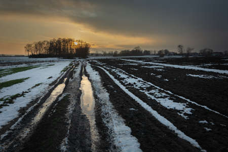 Muddy dirt road, snow in the field, cloud and sunsetの写真素材