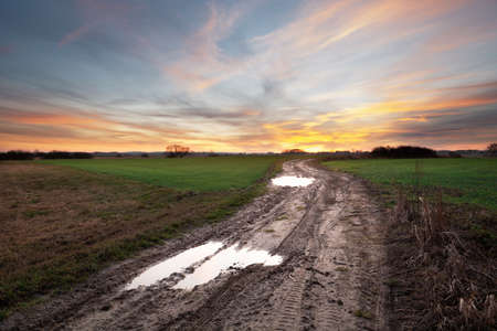 Puddles on a dirt road through fields and beautiful colorful clouds on evening sky