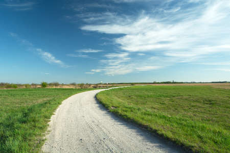 A bend on a gravel road and green meadows, white clouds on a blue sky, spring view