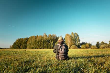 A man with a backpack sitting on the meadow, forest on the horizon and blue skyの写真素材