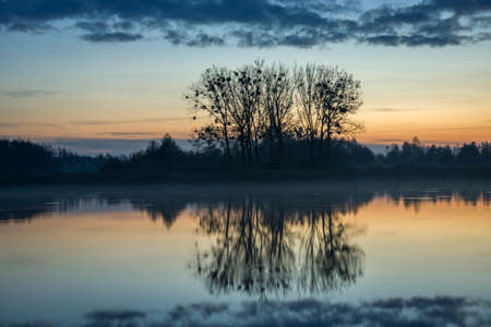 Trees on the shores of a calm lake, fog and clouds