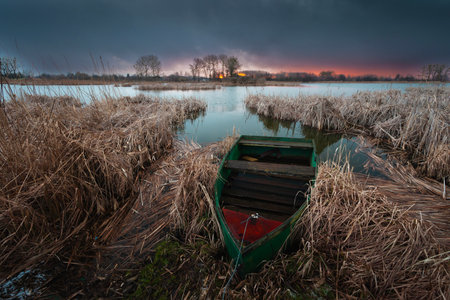 A view of an old fishing boat moored in the dry reeds on the shore of a lake during a cloudy sunset.の写真素材