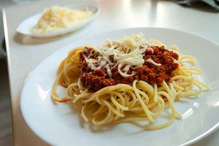 A dish of spaghetti pasta with minced meat in tomato sauce sprinkled with yellow cheese served on a white round big plate stands on the kitchen counterの写真素材
