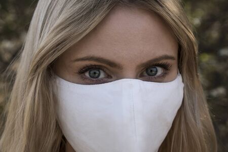 Young blonde girl in a protective mask for spreading of disease virus SARS-CoV-2. Epidemic. Coronavirus. Covid-19. Trees on the background.の写真素材