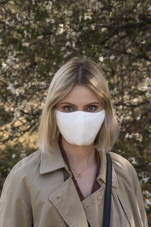 Young blonde girl in a protective mask for spreading of disease virus SARS-CoV-2. Epidemic. Coronavirus. Covid-19. Trees on the background.の写真素材