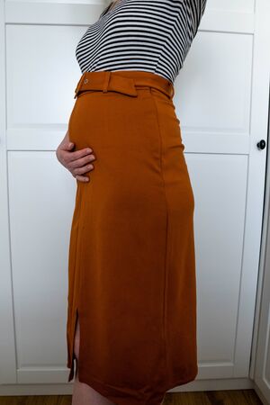 Attractive pregnant young woman in an orange skirt at home and holding her belly. Motherhood, pregnancy, happiness.の写真素材