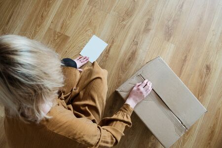 A young woman who is a customer prepares a package for shipping using tape and scissors. Girl consumer holding cardboard box on the floor.の写真素材