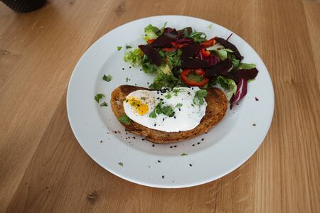 Poached egg on a piece of bread with green salad on a plate and on the wooden table. Healthy Breakfastの写真素材