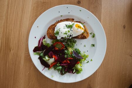 Poached egg on a piece of bread with green salad on a plate and on the wooden table. Healthy Breakfastの写真素材