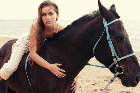 fashion outdoor photo of beautiful young woman with long dark hair and evening makeup posing with black horse,walking by autumn beachの写真素材