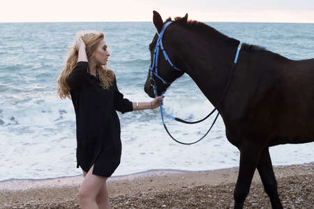 fashion outdoor photo of beautiful young woman with long blond hair  posing with black horse,walking by autumn beachの写真素材