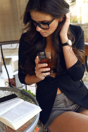 Fashion photo of beautiful sexy business woman with dark curly hair wearing black jacket,watch and  glasses, sitting at cafe and drinking teaの写真素材