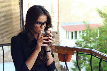 Fashion photo of beautiful sexy business woman with dark curly hair wearing black jacket,watch, glasses and sitting at cafe, drinking teaの写真素材