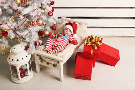holiday photo of cute little Santa baby posing beside Christmas tree at cozy home with New Year decoration and big presentsの写真素材