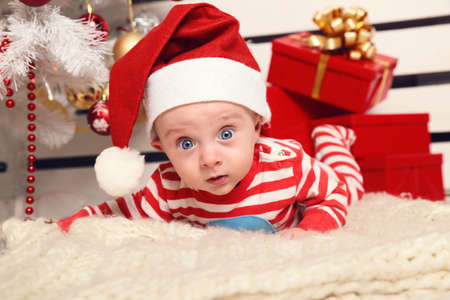 holiday photo of cute little Santa baby posing beside Christmas tree at cozy home with New Year decoration and big presentsの写真素材