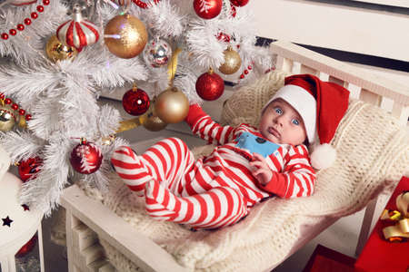 holiday photo of cute little Santa baby posing beside Christmas tree at cozy home with New Year decorationの写真素材