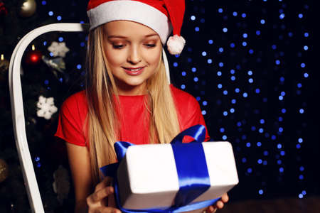 beautiful little girl in santa hat and red dress smiling and holding a box with Christmas present on...