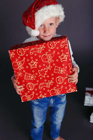 holiday photo with  little boy in santa hat and jeans and a white sweater smiles and holding a box...