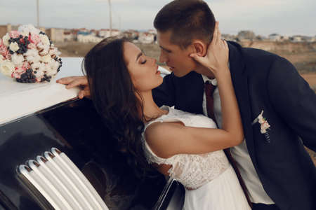fashion outdoor love story, beautiful groom and bride in wedding clothes posing beside a carの写真素材