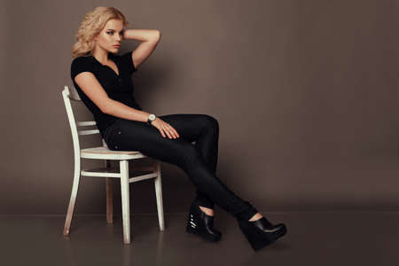 fashion studio photo of beautiful woman with blond curly hair wears casual clothes, sitting on chair
