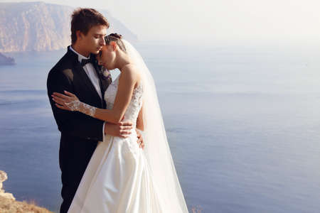 fashion outdoor photo of beautiful couple. gorgeous bride in wedding dress posing with elegant groom on sea costの写真素材