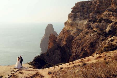 fashion outdoor photo of beautiful couple. gorgeous bride in wedding dress posing with elegant groom on sea costの写真素材