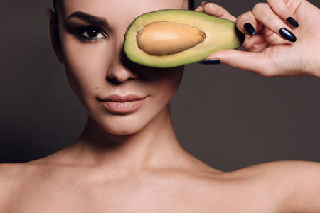 fashion interior photo of gorgeous woman with dark hair and evening makeup, holding avocado in hand