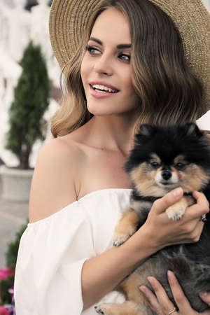 fashion outdoor photo of gorgeous girl with dark hair in elegant clothes, holding small cute puppyの写真素材