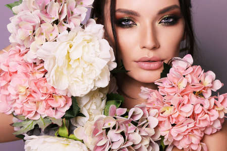 fashion photo of beautiful girl with dark hair in elegant outfit posing among spring flowers in studioの写真素材
