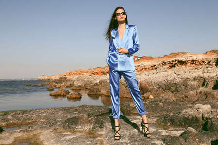 fashion outdoor photo of beautiful woman with dark hair in elegant blue suit posing among stones on the summer beachの写真素材