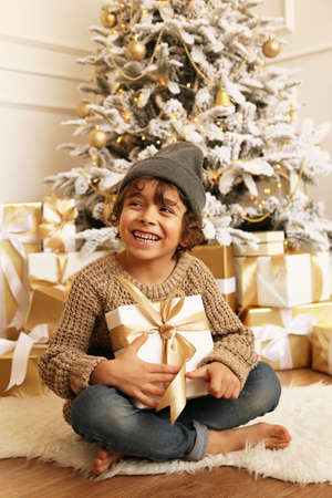 cozy photo of cute little boy with dark hair in cozy clothes posing near decorated Christmas tree with presentsの写真素材