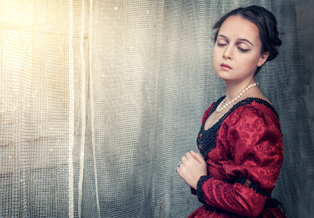 Sad beautiful young woman in red medieval dress near windowの写真素材