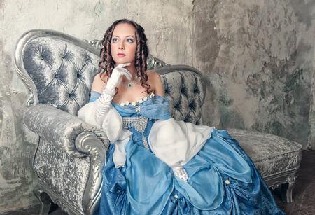 Beautiful young woman in blue medieval dress on the sofaの写真素材