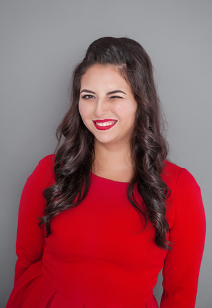 Happy plus size woman in red dress winking on gray backgroundの写真素材