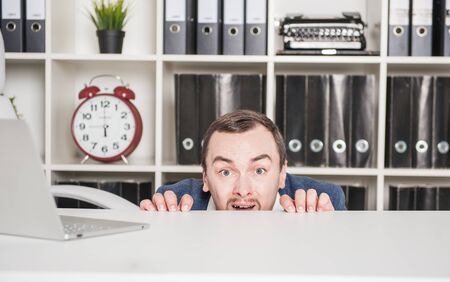 Scared business man hiding behind the table in office