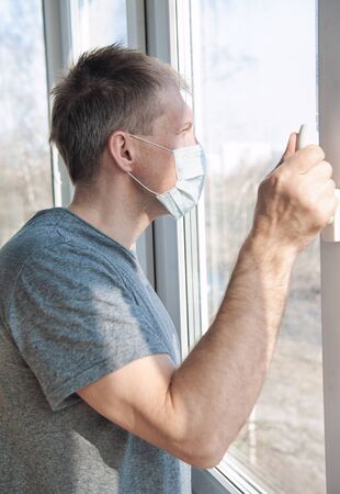 Sick sad man in medical protective face mask looking through window. Coronavirus pandemic covid-19 and quarantine concept
の写真素材