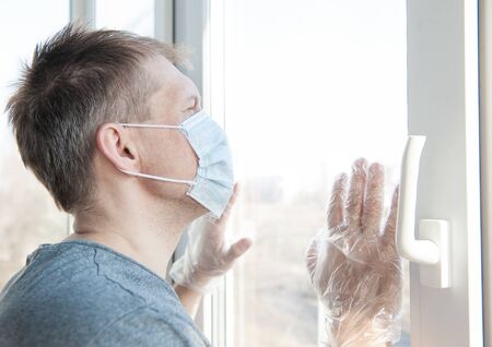 Sick sad man in medical protective face mask looking through window. Coronavirus pandemic covid-19 and quarantine concept
の写真素材