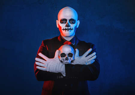 Bold man with scary Halloween make up dead day calavera style holding skull on blue background