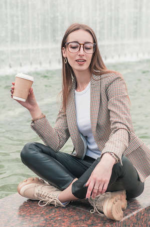 Beautiful fashion stylish woman with cup of coffee sitting on the parapet near fountain background
