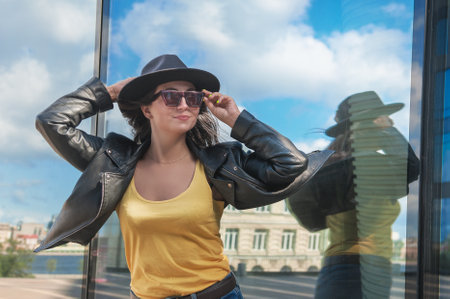 Beautiful young woman with hat and sunglasses near the mirror wall of city buildingの写真素材