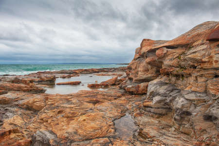Big boulders of red rock and sea waves against the dramatic blue sky backgroundの写真素材